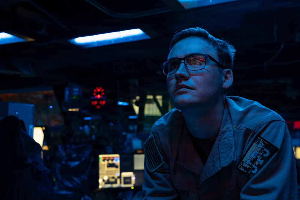 A military personnel member wearing a uniform with a U.S. flag patch and a ship/unit insignia patch is stationed at what appears to be a naval vessel's combat information center or bridge. The individual is wearing glasses and gazing upward or forward in a dimly lit operational environment. The scene is illuminated by blue ambient lighting and red indicator lights, consistent with a low-light tactical setting.