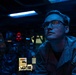 A military personnel member wearing a uniform with a U.S. flag patch and a ship/unit insignia patch is stationed at what appears to be a naval vessel's combat information center or bridge. The individual is wearing glasses and gazing upward or forward in a dimly lit operational environment. The scene is illuminated by blue ambient lighting and red indicator lights, consistent with a low-light tactical setting.