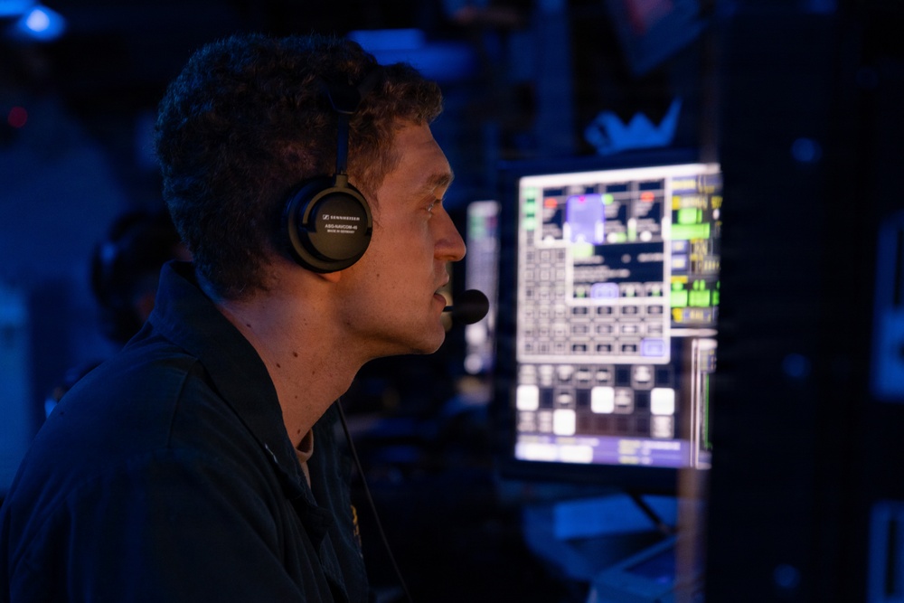 A male operator wearing a Sennheiser headset with boom microphone is seated at a workstation in a dimly lit control room with blue ambient lighting. He is viewed in profile, monitoring multiple screens displaying what appear to be tactical or operational system interfaces with green and white data readouts. The environment suggests a military or naval command and control setting.