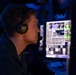 A male operator wearing a Sennheiser headset with boom microphone is seated at a workstation in a dimly lit control room with blue ambient lighting. He is viewed in profile, monitoring multiple screens displaying what appear to be tactical or operational system interfaces with green and white data readouts. The environment suggests a military or naval command and control setting.