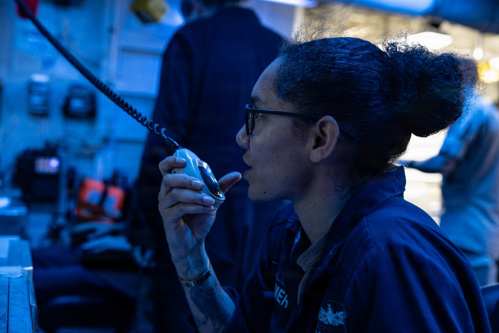 A female military or naval personnel member is speaking into a sound-powered telephone or shipboard intercom handset in what appears to be a ship's combat information center or bridge. The environment is illuminated with blue tactical lighting, characteristic of naval vessel operations. She is wearing a navy blue uniform and glasses, with her hair pulled up.