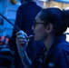 A female military or naval personnel member is speaking into a sound-powered telephone or shipboard intercom handset in what appears to be a ship's combat information center or bridge. The environment is illuminated with blue tactical lighting, characteristic of naval vessel operations. She is wearing a navy blue uniform and glasses, with her hair pulled up.