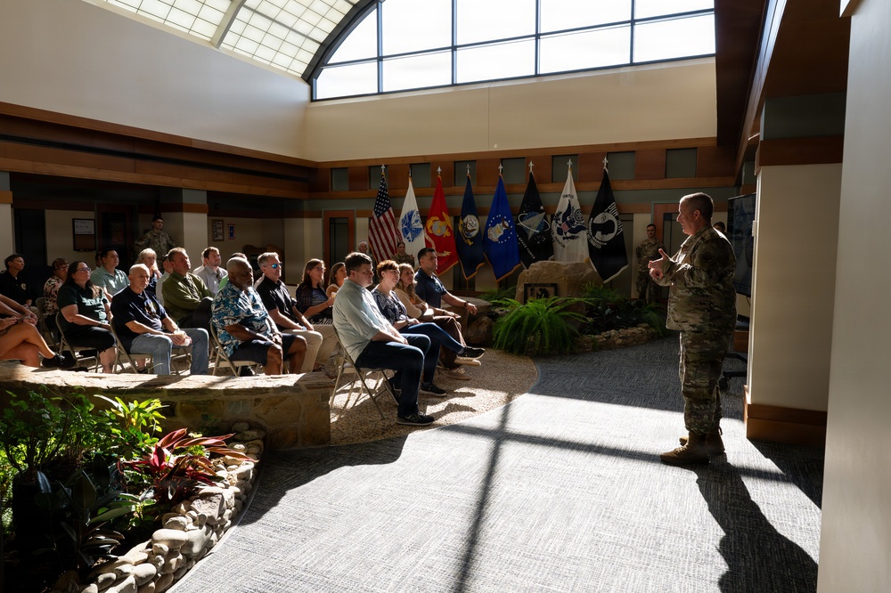 A military officer in camouflage uniform addresses a seated civilian audience in what appears to be a formal military facility lobby or atrium. Multiple military branch flags are displayed in the background, including the U.S. flag, Marine Corps, Air Force, Space Force, and POW/MIA flags. The setting suggests an official briefing, orientation, or community engagement event.