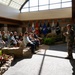 A military officer in camouflage uniform addresses a seated civilian audience in what appears to be a formal military facility lobby or atrium. Multiple military branch flags are displayed in the background, including the U.S. flag, Marine Corps, Air Force, Space Force, and POW/MIA flags. The setting suggests an official briefing, orientation, or community engagement event.