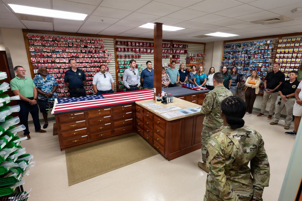 A group of civilians and military personnel are gathered in what appears to be a flag room or flag processing facility, likely at a government or military installation. Two soldiers in Army Combat Uniforms (ACU/OCP pattern) are facing a group of approximately 20 civilians and uniformed personnel, suggesting a briefing or tour is taking place. An American flag is displayed unfolded on a wooden counter/workstation in the center of the room.