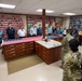 A group of civilians and military personnel are gathered in what appears to be a flag room or flag processing facility, likely at a government or military installation. Two soldiers in Army Combat Uniforms (ACU/OCP pattern) are facing a group of approximately 20 civilians and uniformed personnel, suggesting a briefing or tour is taking place. An American flag is displayed unfolded on a wooden counter/workstation in the center of the room.