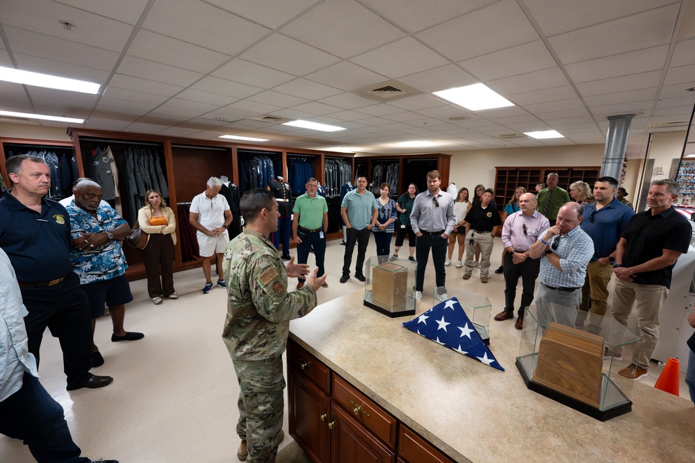 A uniformed military soldier in camouflage (OCP/MultiCam) uniform is briefing a group of approximately 25 civilians in what appears to be a military uniform or mortuary affairs facility. On the counter in front of the speaker are display items including a folded American flag, glass display cases, and wooden boxes, suggesting this may be a dignified transfer or mortuary affairs briefing room. The civilians appear attentive and are gathered in a semicircle around the presenter.