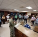 A uniformed military soldier in camouflage (OCP/MultiCam) uniform is briefing a group of approximately 25 civilians in what appears to be a military uniform or mortuary affairs facility. On the counter in front of the speaker are display items including a folded American flag, glass display cases, and wooden boxes, suggesting this may be a dignified transfer or mortuary affairs briefing room. The civilians appear attentive and are gathered in a semicircle around the presenter.
