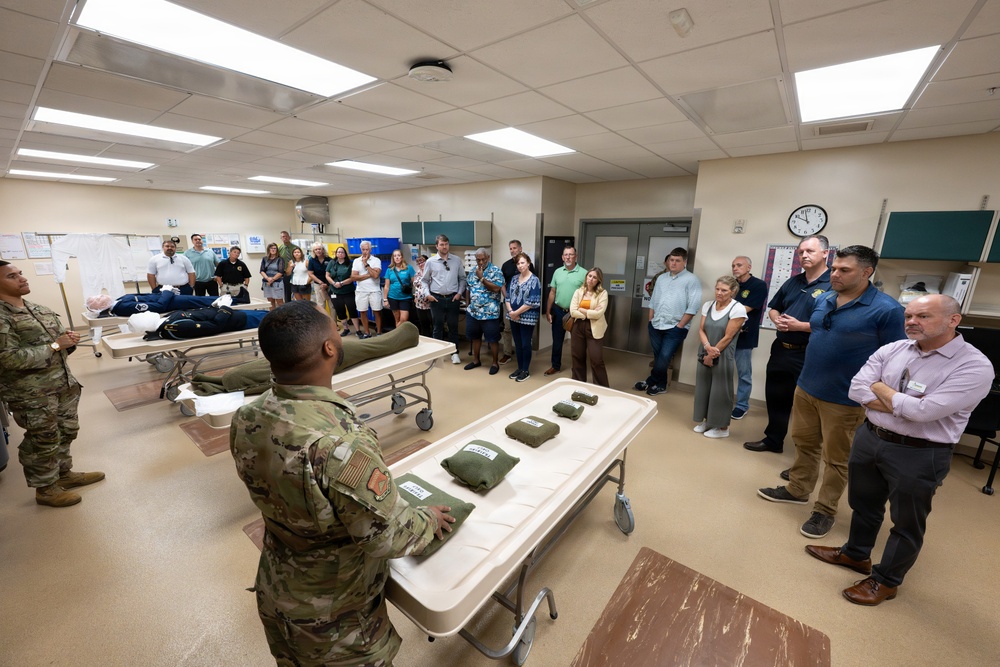 A military briefing or demonstration is taking place in what appears to be a medical training facility. Two soldiers in camouflage uniforms are presenting to a group of approximately 25-30 civilians gathered in a room equipped with medical stretchers/gurneys. Items that appear to be military medical equipment or field gear are displayed on the tables in the foreground.