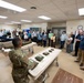A military briefing or demonstration is taking place in what appears to be a medical training facility. Two soldiers in camouflage uniforms are presenting to a group of approximately 25-30 civilians gathered in a room equipped with medical stretchers/gurneys. Items that appear to be military medical equipment or field gear are displayed on the tables in the foreground.