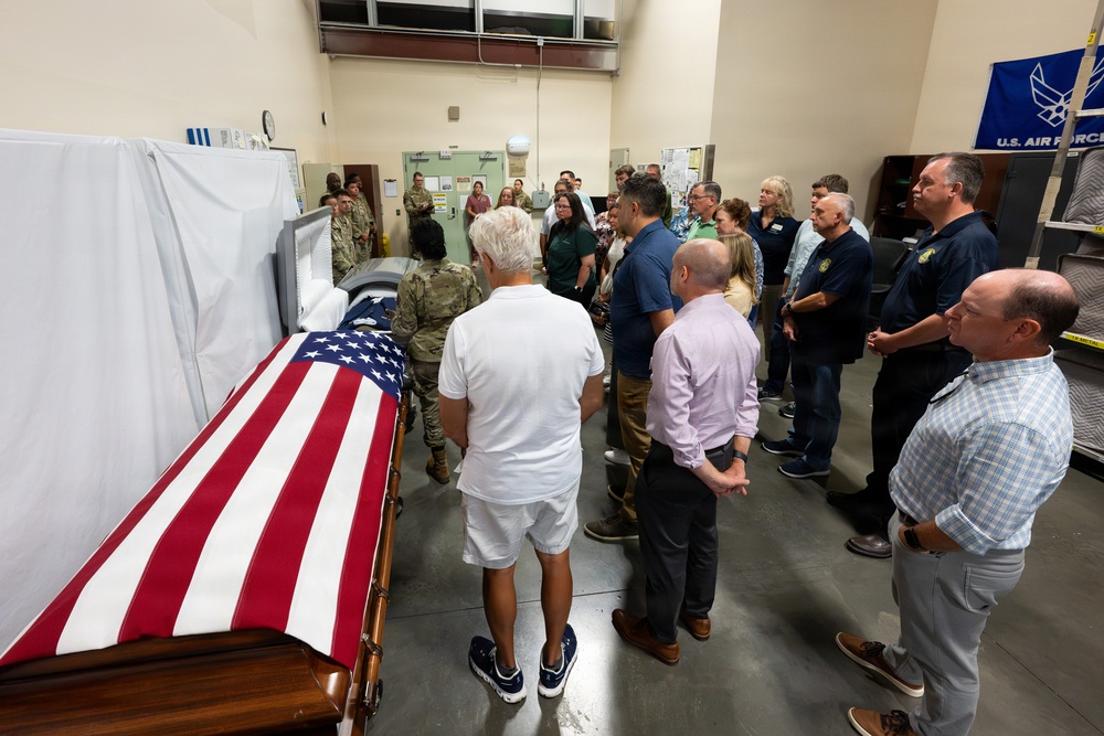 A group of civilians and military personnel are gathered in what appears to be a military mortuary or dignified transfer facility. A flag-draped casket covered with an American flag is prominently visible in the foreground. A U.S. Air Force banner is visible in the background, suggesting this is an official military facility.
