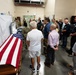 A group of civilians and military personnel are gathered in what appears to be a military mortuary or dignified transfer facility. A flag-draped casket covered with an American flag is prominently visible in the foreground. A U.S. Air Force banner is visible in the background, suggesting this is an official military facility.