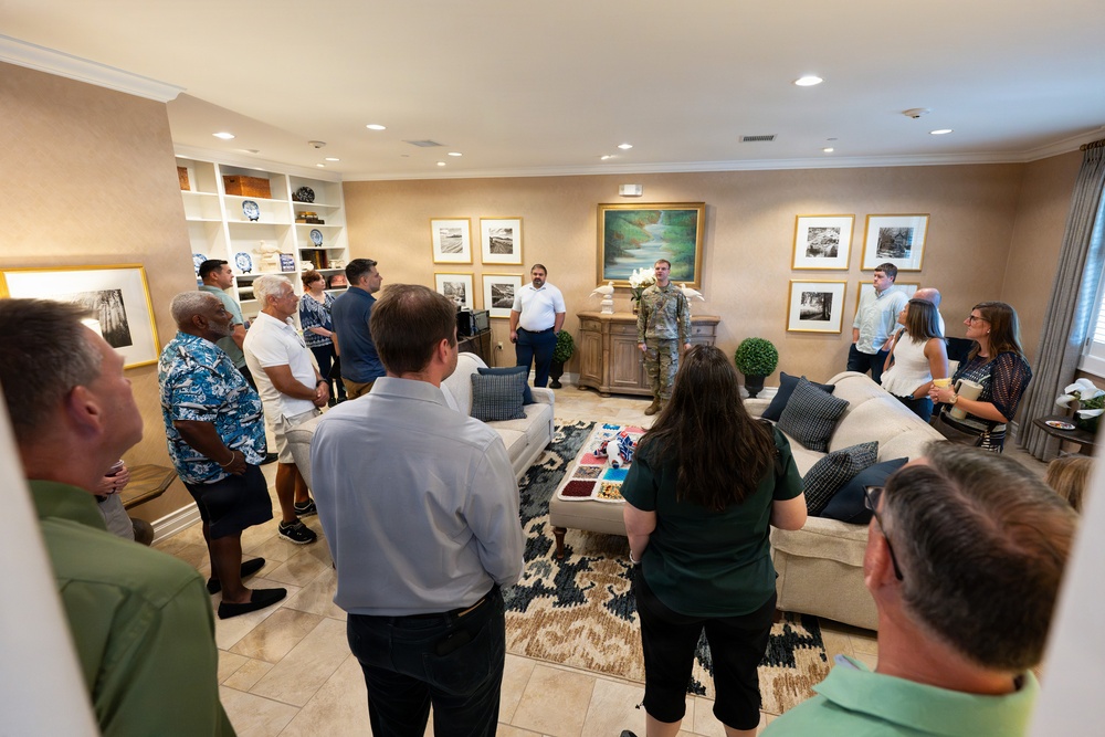 A group of approximately 15-20 people are gathered in a well-appointed living room, listening to a person in military camouflage uniform who appears to be addressing the group. The setting resembles an official residence or upscale quarters, decorated with framed artwork, built-in shelving, and furnished with neutral-toned sofas. The gathering has the appearance of a formal or semi-formal briefing or reception event.