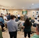 A group of approximately 15-20 people are gathered in a well-appointed living room, listening to a person in military camouflage uniform who appears to be addressing the group. The setting resembles an official residence or upscale quarters, decorated with framed artwork, built-in shelving, and furnished with neutral-toned sofas. The gathering has the appearance of a formal or semi-formal briefing or reception event.
