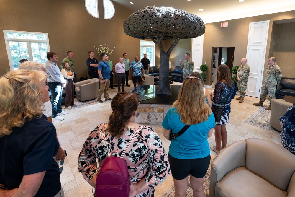 A group of civilians and military personnel in camouflage uniforms are gathered in an upscale interior lobby featuring a large decorative metal tree sculpture as a centerpiece. A military service member appears to be briefing or presenting to the assembled group. The setting suggests an official tour or orientation event at a military facility.