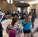 A group of civilians and military personnel in camouflage uniforms are gathered in an upscale interior lobby featuring a large decorative metal tree sculpture as a centerpiece. A military service member appears to be briefing or presenting to the assembled group. The setting suggests an official tour or orientation event at a military facility.