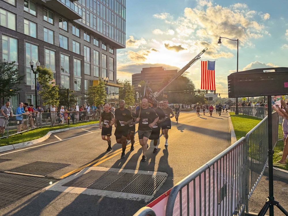 A group of runners dressed in matching black shirts and shorts participate in what appears to be a patriotic road race or memorial run in an urban setting. A large American flag is prominently displayed, suspended from a fire truck ladder boom in the background. Spectators line metal barricades along the course as the event takes place during golden hour sunset lighting.