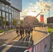 A group of runners dressed in matching black shirts and shorts participate in what appears to be a patriotic road race or memorial run in an urban setting. A large American flag is prominently displayed, suspended from a fire truck ladder boom in the background. Spectators line metal barricades along the course as the event takes place during golden hour sunset lighting.