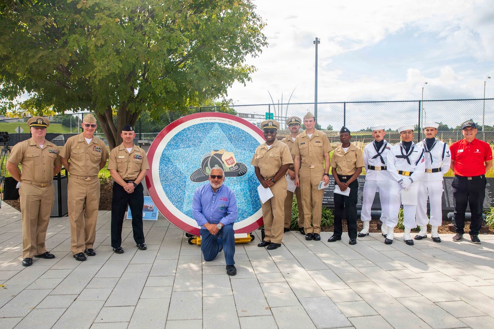 A group of approximately 12 individuals, primarily U.S. Navy personnel in various uniforms, pose for a formal group photograph outdoors. They are gathered around a large circular mosaic artwork featuring a blue star design with a firefighter helmet and the number '911' at its center. The setting appears to be a memorial or dedication ceremony at an outdoor plaza.