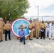 A group of approximately 12 individuals, primarily U.S. Navy personnel in various uniforms, pose for a formal group photograph outdoors. They are gathered around a large circular mosaic artwork featuring a blue star design with a firefighter helmet and the number '911' at its center. The setting appears to be a memorial or dedication ceremony at an outdoor plaza.