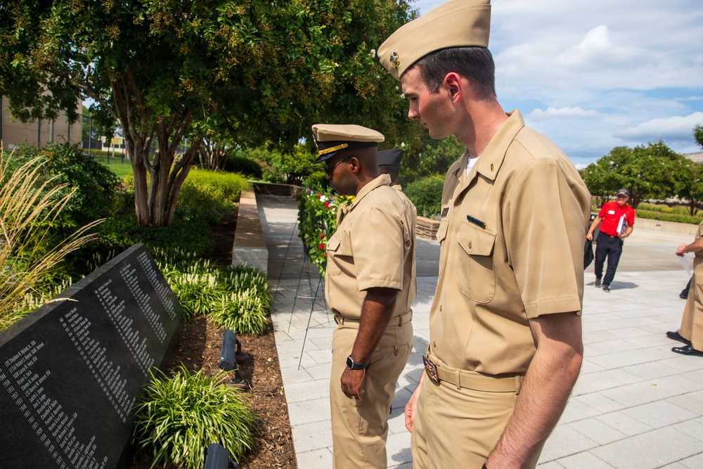 Two U.S. Navy personnel in service khaki uniforms stand solemnly before a dark granite memorial wall engraved with names. A floral wreath is visible in the background near the memorial, suggesting a commemorative ceremony is taking place. The setting appears to be an outdoor memorial garden at a military installation.