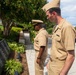 Two U.S. Navy personnel in service khaki uniforms stand solemnly before a dark granite memorial wall engraved with names. A floral wreath is visible in the background near the memorial, suggesting a commemorative ceremony is taking place. The setting appears to be an outdoor memorial garden at a military installation.