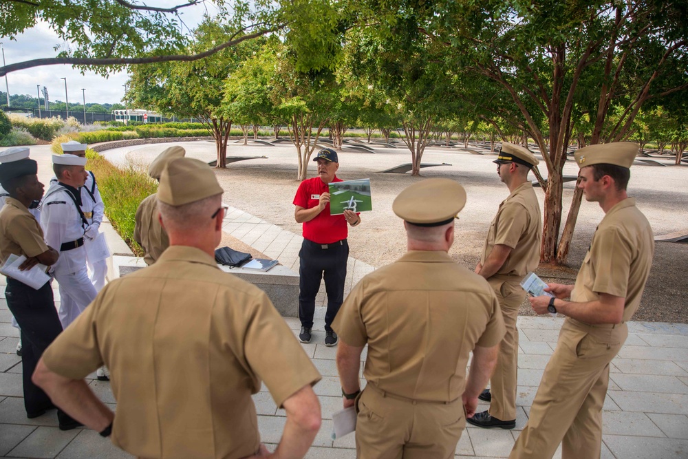 A civilian guide in a red shirt is conducting a tour for a group of U.S. Navy personnel at what appears to be the Pentagon Memorial in Arlington, Virginia. The guide is holding up a photograph or diagram while addressing the uniformed service members gathered around him. The setting features the memorial's distinctive cantilevered benches and crape myrtle trees arranged across the paved grounds.
