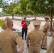 A civilian guide in a red shirt is conducting a tour for a group of U.S. Navy personnel at what appears to be the Pentagon Memorial in Arlington, Virginia. The guide is holding up a photograph or diagram while addressing the uniformed service members gathered around him. The setting features the memorial's distinctive cantilevered benches and crape myrtle trees arranged across the paved grounds.