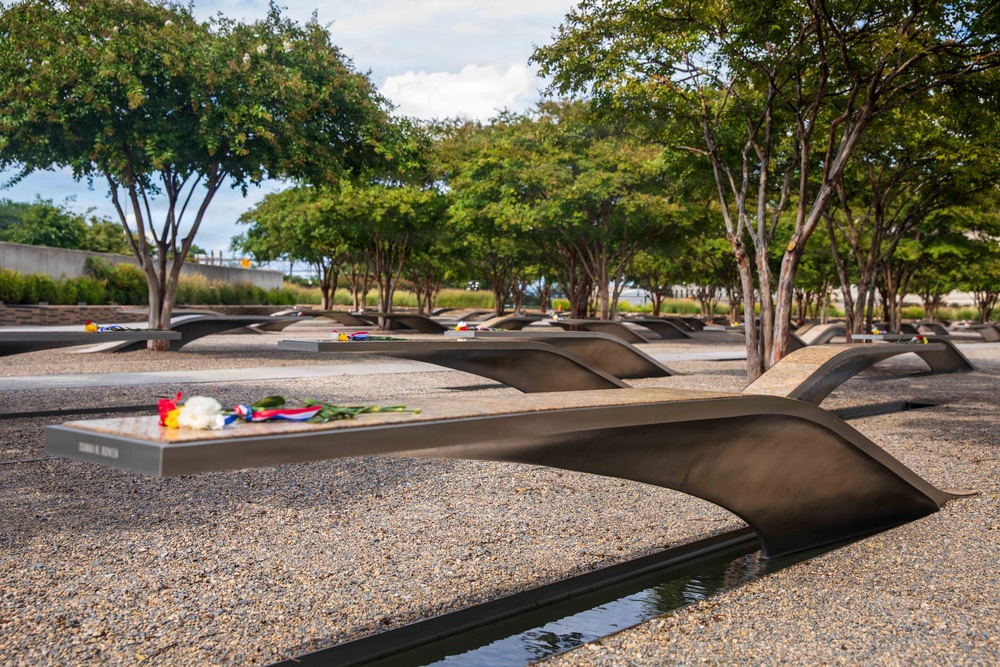 The image depicts an outdoor memorial site featuring multiple curved, cantilevered bench-like structures made of dark metal, each bearing an engraved name and containing a small reflecting pool beneath. Flowers, ribbons, and small tributes have been placed on several of the memorial benches. The setting includes gravel ground cover, mature trees, and a clear sky, consistent with the Pentagon Memorial in Arlington, Virginia.