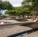 The image depicts an outdoor memorial site featuring multiple curved, cantilevered bench-like structures made of dark metal, each bearing an engraved name and containing a small reflecting pool beneath. Flowers, ribbons, and small tributes have been placed on several of the memorial benches. The setting includes gravel ground cover, mature trees, and a clear sky, consistent with the Pentagon Memorial in Arlington, Virginia.