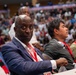 A man in a navy blue blazer and white shirt wearing a red lanyard credential is seated in what appears to be a large indoor arena or sports venue. He is looking directly at the camera while surrounded by other attendees. The venue appears to be hosting a large event with a packed crowd visible in the background.