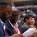 The image shows a formal gathering or conference with attendees seated at tables in what appears to be a large venue or auditorium. In the foreground, a man wearing glasses, a blue blazer, and a red tie is focused on reading printed documents. The background shows a crowded audience in tiered seating, suggesting a large-scale event.