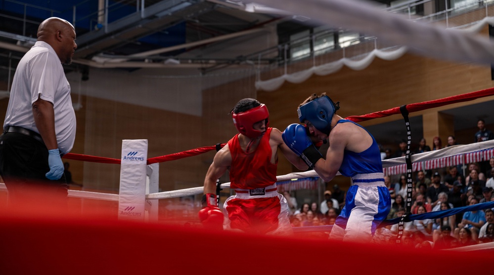 Two amateur boxers are competing in a boxing ring, with one wearing red gear and the other wearing blue gear. A referee in a white shirt and black pants observes the bout closely from the corner. The match appears to be taking place in an indoor venue with a sizeable crowd watching from the stands.