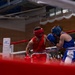 Two amateur boxers are competing in a boxing ring, with one wearing red gear and the other wearing blue gear. A referee in a white shirt and black pants observes the bout closely from the corner. The match appears to be taking place in an indoor venue with a sizeable crowd watching from the stands.