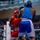Two amateur boxers compete in a boxing match inside an indoor arena with a visible audience in the background. One boxer wears red gear (headgear, gloves, shorts, and tank top) while the opponent wears blue gear. The match appears to be an organized amateur event, evidenced by the sponsor banners on the ring corner padding.