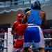 Two amateur boxers compete in a boxing match inside an indoor arena with a visible audience in the background. One boxer wears red gear (headgear, gloves, shorts, and tank top) while the opponent wears blue gear. The match appears to be an organized amateur event, evidenced by the sponsor banners on the ring corner padding.