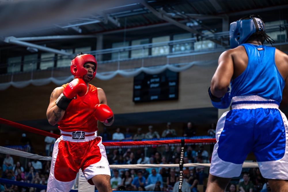 Two amateur boxers compete in a boxing ring during what appears to be a sanctioned bout in an indoor arena. The boxer in red (wearing Ringside brand gear) faces off against an opponent in blue, both wearing protective headgear and mouthguards. A large crowd of spectators fills the background seating area.