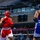 Two amateur boxers compete in a boxing ring during what appears to be a sanctioned bout in an indoor arena. The boxer in red (wearing Ringside brand gear) faces off against an opponent in blue, both wearing protective headgear and mouthguards. A large crowd of spectators fills the background seating area.