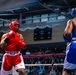 Two amateur boxers compete in a boxing ring during what appears to be a sanctioned bout in an indoor arena. The boxer in red (wearing Ringside brand gear) faces off against an opponent in blue, both wearing protective headgear and mouthguards. A large crowd of spectators fills the background seating area.