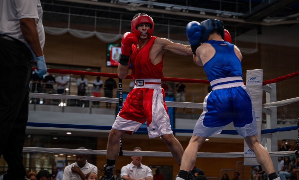 Two amateur boxers are competing in a boxing ring, with one fighter wearing red gear and the other in blue gear. The red-corner fighter appears to be throwing or landing a punch while the blue-corner fighter is in a defensive or receiving position. The match is taking place in an indoor venue with spectators visible in the background.