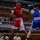 Two amateur boxers are competing in a boxing ring, with one fighter wearing red gear and the other in blue gear. The red-corner fighter appears to be throwing or landing a punch while the blue-corner fighter is in a defensive or receiving position. The match is taking place in an indoor venue with spectators visible in the background.