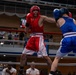Two amateur boxers are competing in a boxing ring, with one fighter wearing red gear and the other in blue gear. The red-corner fighter appears to be throwing or landing a punch while the blue-corner fighter is in a defensive or receiving position. The match is taking place in an indoor venue with spectators visible in the background.