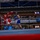 An amateur boxing match is taking place inside an indoor arena with spectators watching from the stands and ringside. Two boxers — one in red gear and one in blue gear — are actively engaged in a bout near a corner of the ring, with a referee observing closely. The venue features visible sponsor signage including 'Andrews' and 'MWR', suggesting a military or institutional event.