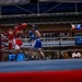 An amateur boxing match is taking place inside an indoor arena with spectators watching from the stands and ringside. Two boxers — one in red gear and one in blue gear — are actively engaged in a bout near a corner of the ring, with a referee observing closely. The venue features visible sponsor signage including 'Andrews' and 'MWR', suggesting a military or institutional event.