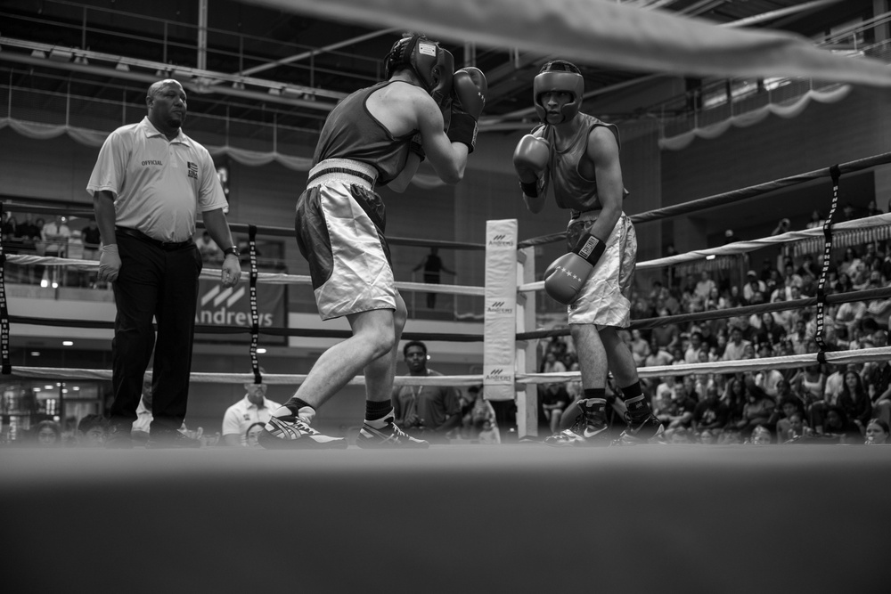 A black-and-white photograph captures an amateur boxing match in progress inside a well-attended arena. Two boxers wearing headgear and gloves are engaged in close-range combat in the ring, while a referee in an 'Official' shirt observes from the side. The venue appears to be a multi-level indoor facility with a large crowd of spectators visible in the background.