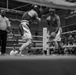 A black-and-white photograph captures an amateur boxing match in progress inside a well-attended arena. Two boxers wearing headgear and gloves are engaged in close-range combat in the ring, while a referee in an 'Official' shirt observes from the side. The venue appears to be a multi-level indoor facility with a large crowd of spectators visible in the background.