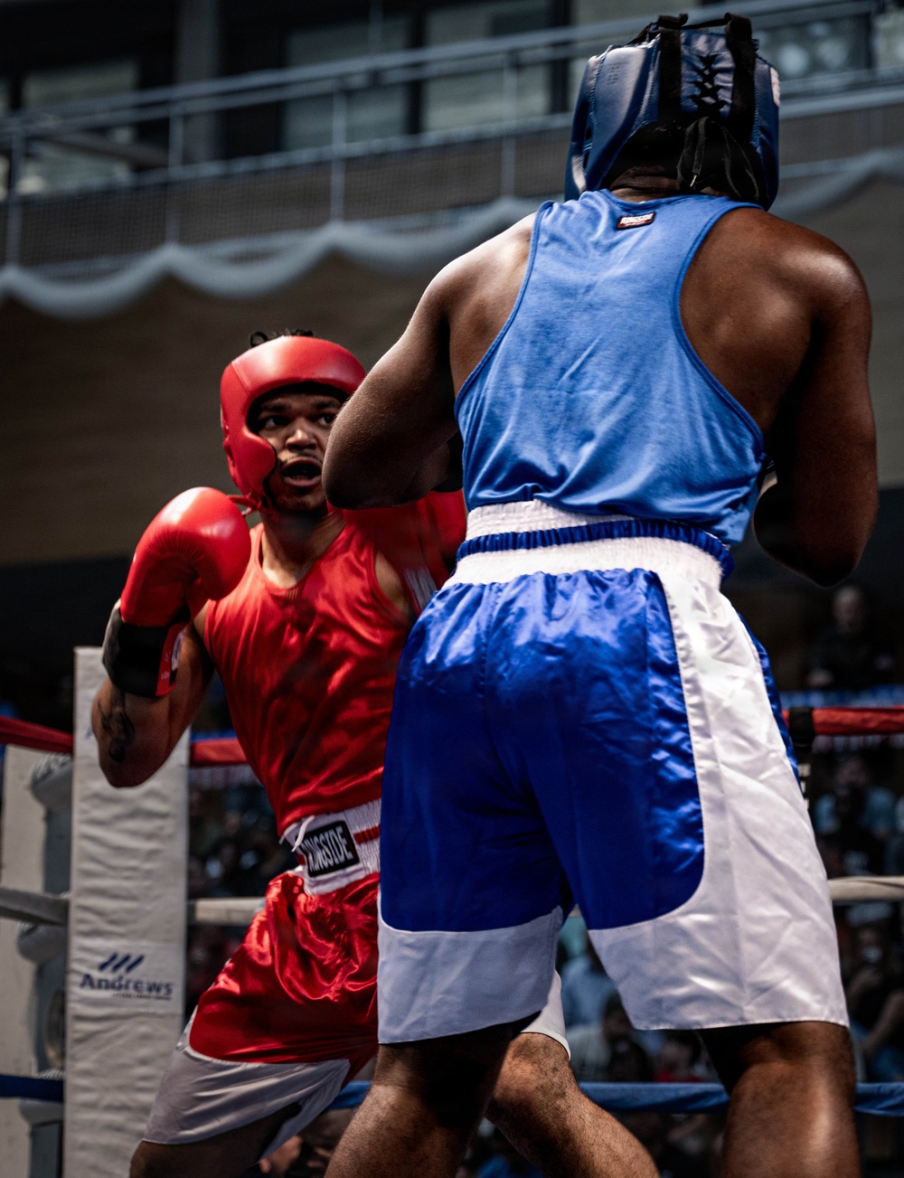 Two amateur boxers are engaged in an active bout inside a boxing ring. The fighter in red is on the defensive, absorbing or slipping a punch from the fighter in blue. Both competitors are wearing full amateur-style headgear and matching colored gloves and shorts.