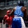 Two amateur boxers are engaged in an active bout inside a boxing ring. The fighter in red is on the defensive, absorbing or slipping a punch from the fighter in blue. Both competitors are wearing full amateur-style headgear and matching colored gloves and shorts.