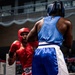 Two amateur boxers are engaged in an active bout inside a boxing ring. The fighter in red is on the defensive, absorbing or slipping a punch from the fighter in blue. Both competitors are wearing full amateur-style headgear and matching colored gloves and shorts.