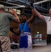 A young boxer in blue and white 'Ringside' shorts has his arm raised by a referee in a white USA Boxing shirt, signaling a victory. The scene takes place inside a boxing ring with a crowd of spectators visible in the background. A third person in a green shirt, likely a coach or corner man, is also present and appears to be congratulating the winner.